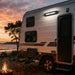 Camper trailer parked by a campfire with a sunset over water
