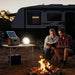 Two people sitting by a campfire with a solar panel and portable light in front of an RV.