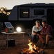 Two people sitting by a campfire with a solar panel and portable light in front of an RV.