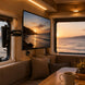 Interior of a vehicle with a TV displaying a sunset over water, a couch, and a coffee cup on a table.
